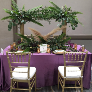 Purple-covered banquet table with tropical greenery and gold chairs in a decorated indoor venue