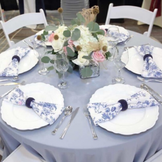Round table with blue tablecloth, white plates, silverware, and a pastel floral centerpiece.
