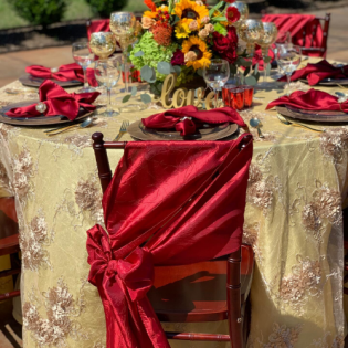 Elegant outdoor table set with red satin chair covers, gold tablecloth, and a colorful floral centerpiece