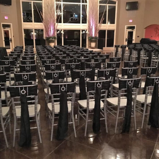Rows of black chairs with white cushions arranged in a banquet hall before tall windows.