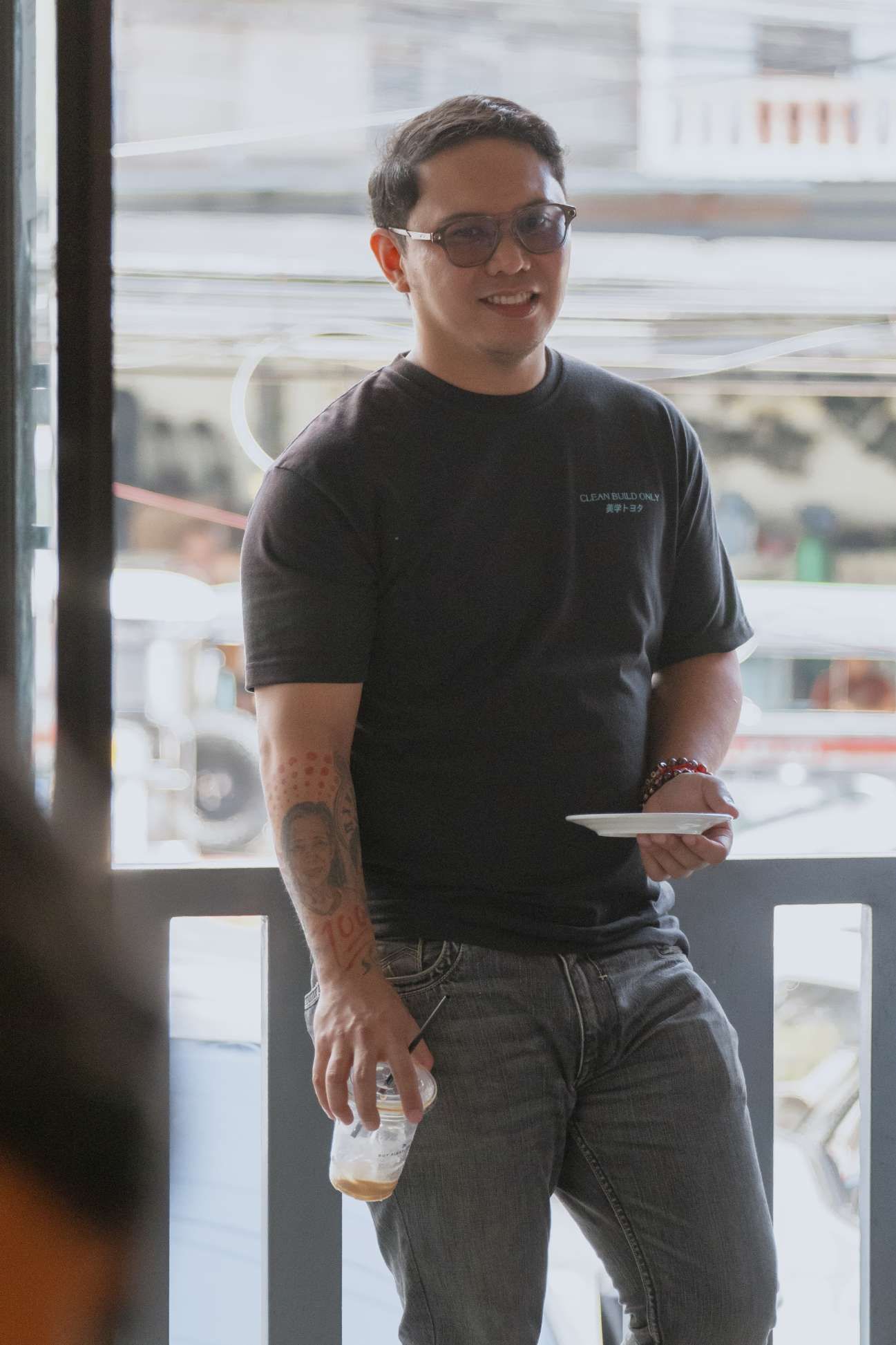 A man wearing sunglasses and a black shirt is standing in front of a window holding a cup of coffee.