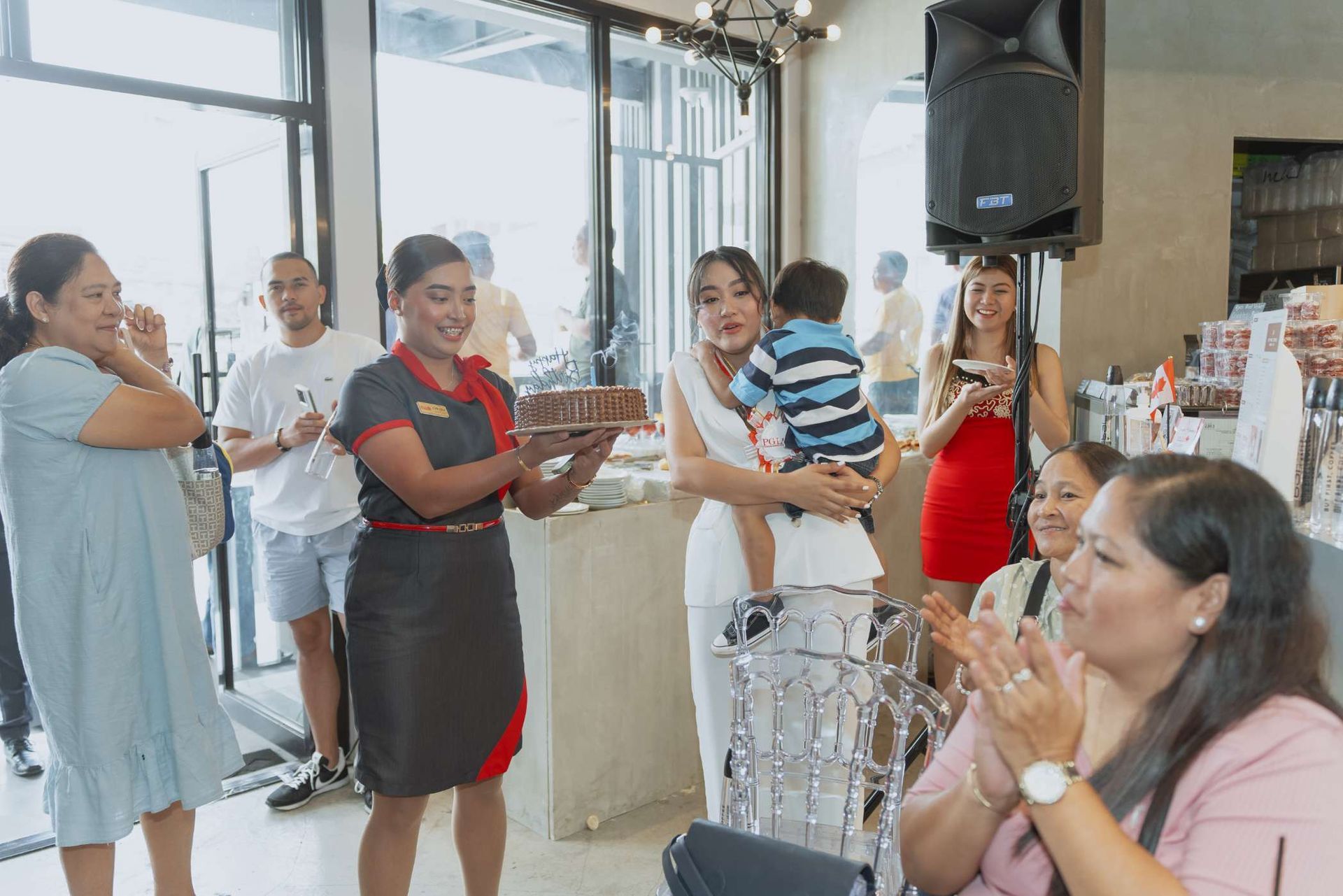 A group of people are standing around a woman holding a baby and a cake.