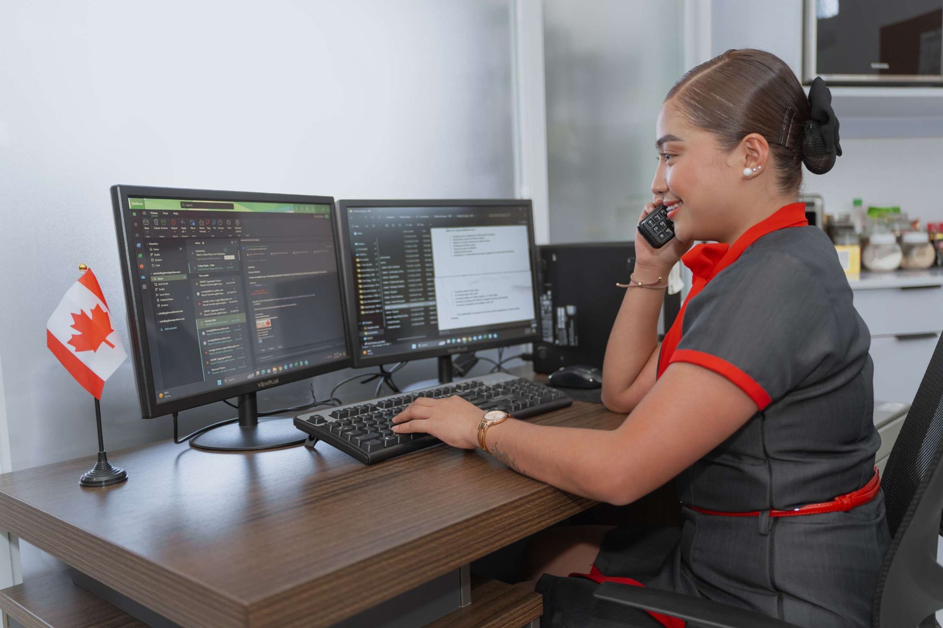 A woman is sitting at a desk talking on a cell phone while using a computer.