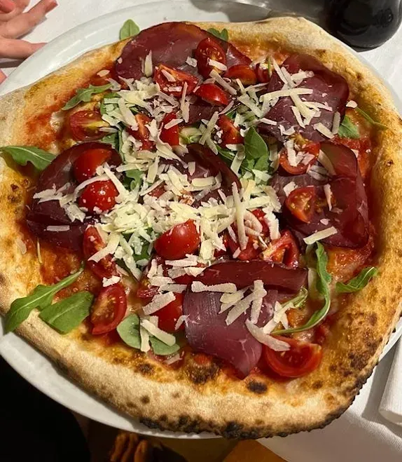 Una pizza a crosta sottile condita con bresaola, pomodorini, rucola e scaglie di parmigiano, servita su un piatto bianco.