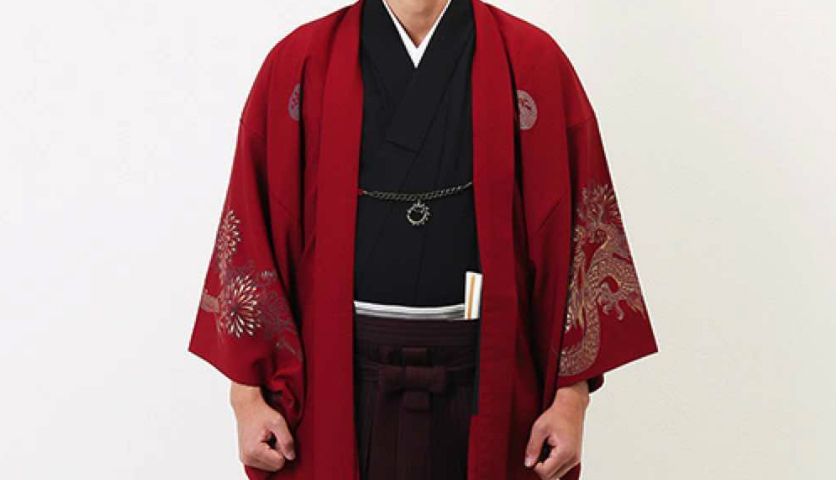 Persona que viste una túnica roja estilo kimono sobre un atuendo negro, de pie frente a un fondo liso.