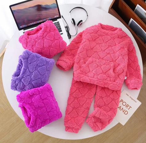 Conjunto infantil rosa de forro polar con tres camisetas dobladas a juego y un ordenador portátil sobre una mesa.