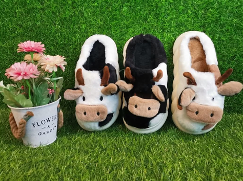 Tres pantuflas de vaca de peluche sobre la hierba junto a una pequeña maceta con flores rosas.