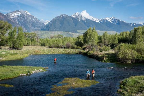 Two people are fishing in a river with mountains in the background.