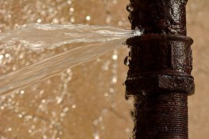 Leaking rusty pipe sprays water against a beige wall.