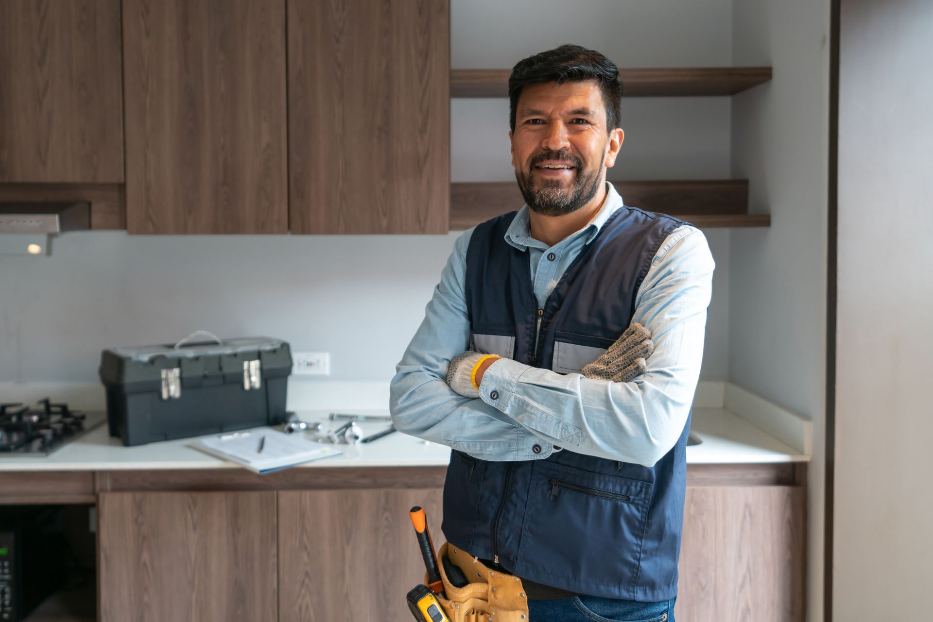 Smiling handyman with arms crossed in a kitchen, wearing a tool belt. Dark cabinets and toolbox are visible.