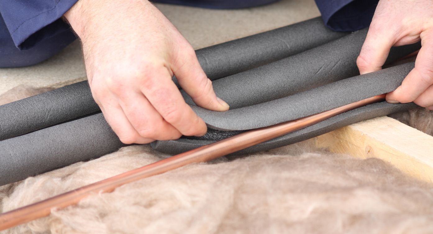 Hands installing black foam pipe insulation over copper pipes in a wooden structure.