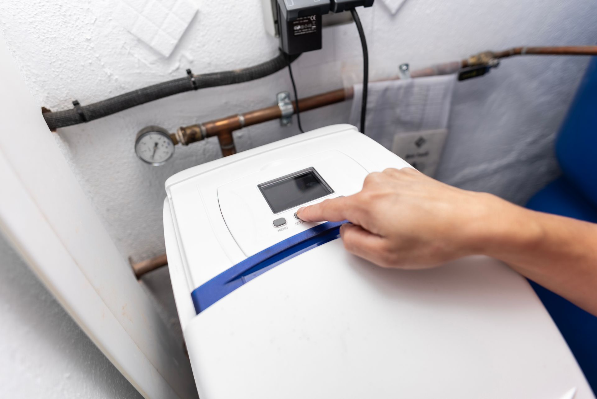 Hand pressing a button on a white water softener unit attached to pipes, indoors.
