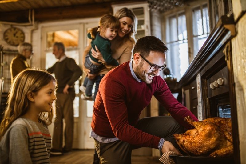 Family gathered around a man taking a turkey from an oven. Child watches, others in background. Warm setting.