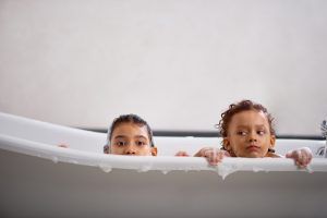 Two young girls in a white bathtub, peeking over the edge, with suds.