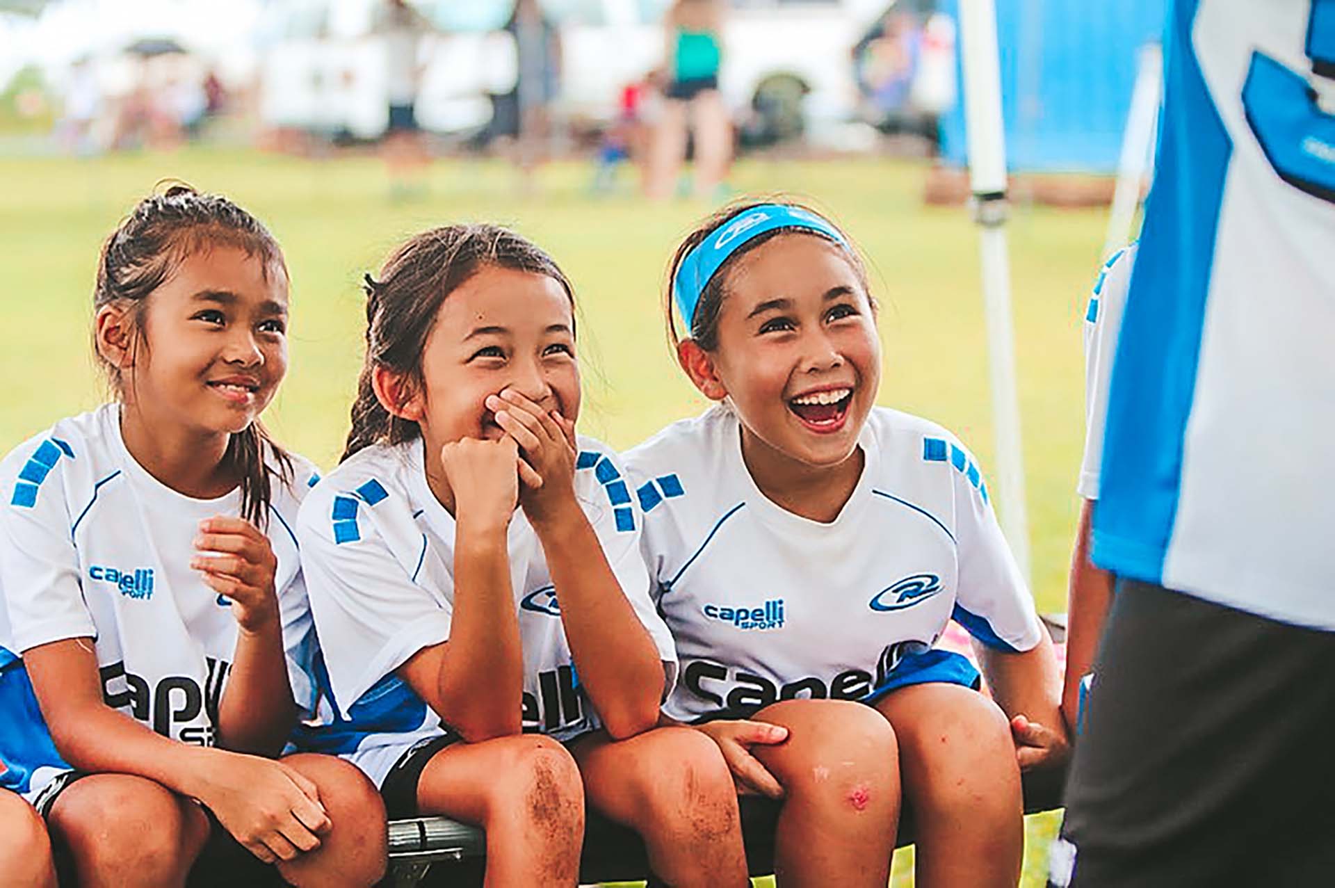 Three children in white sports uniforms sit on a bench, smiling and watching an event.