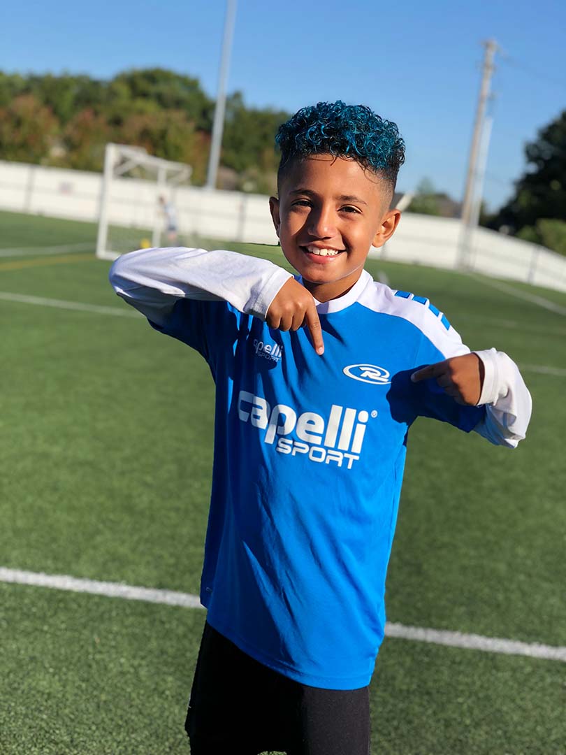 Young soccer player in a blue jersey smiling on a field, pointing to his shirt.