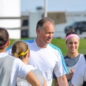 Coach talking with athletes on a sports field, with players in white jerseys and pink headbands.
