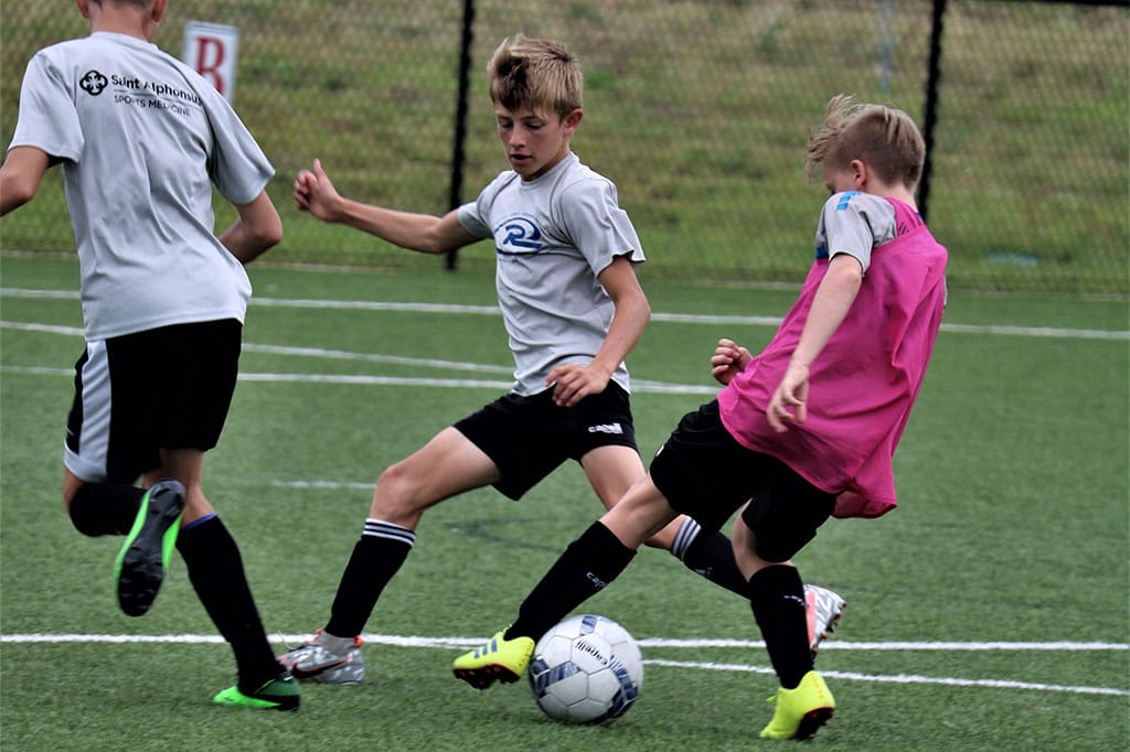 Youth soccer players contest the ball on a field, with one in a pink bib tackling a player in gray.