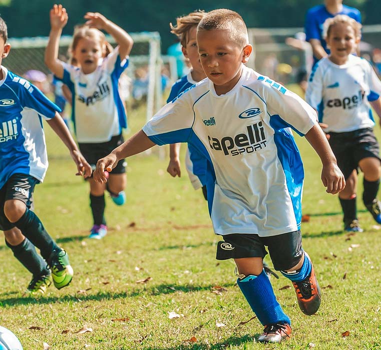 Children playing soccer on a grassy field, with one boy in a white and blue jersey running toward the ball.