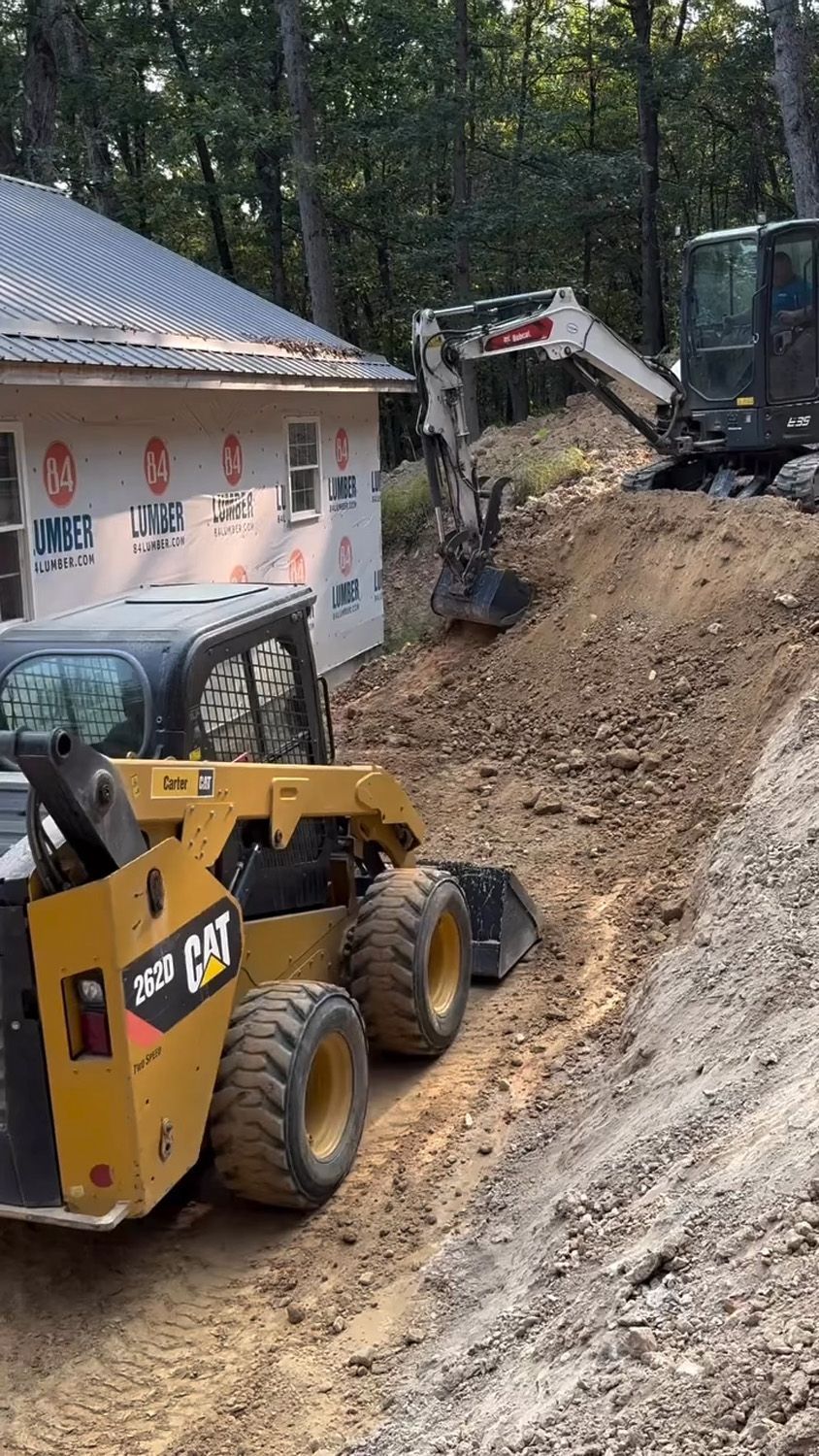 Yellow construction loader and excavator on a dirt hillside beside a house under construction.