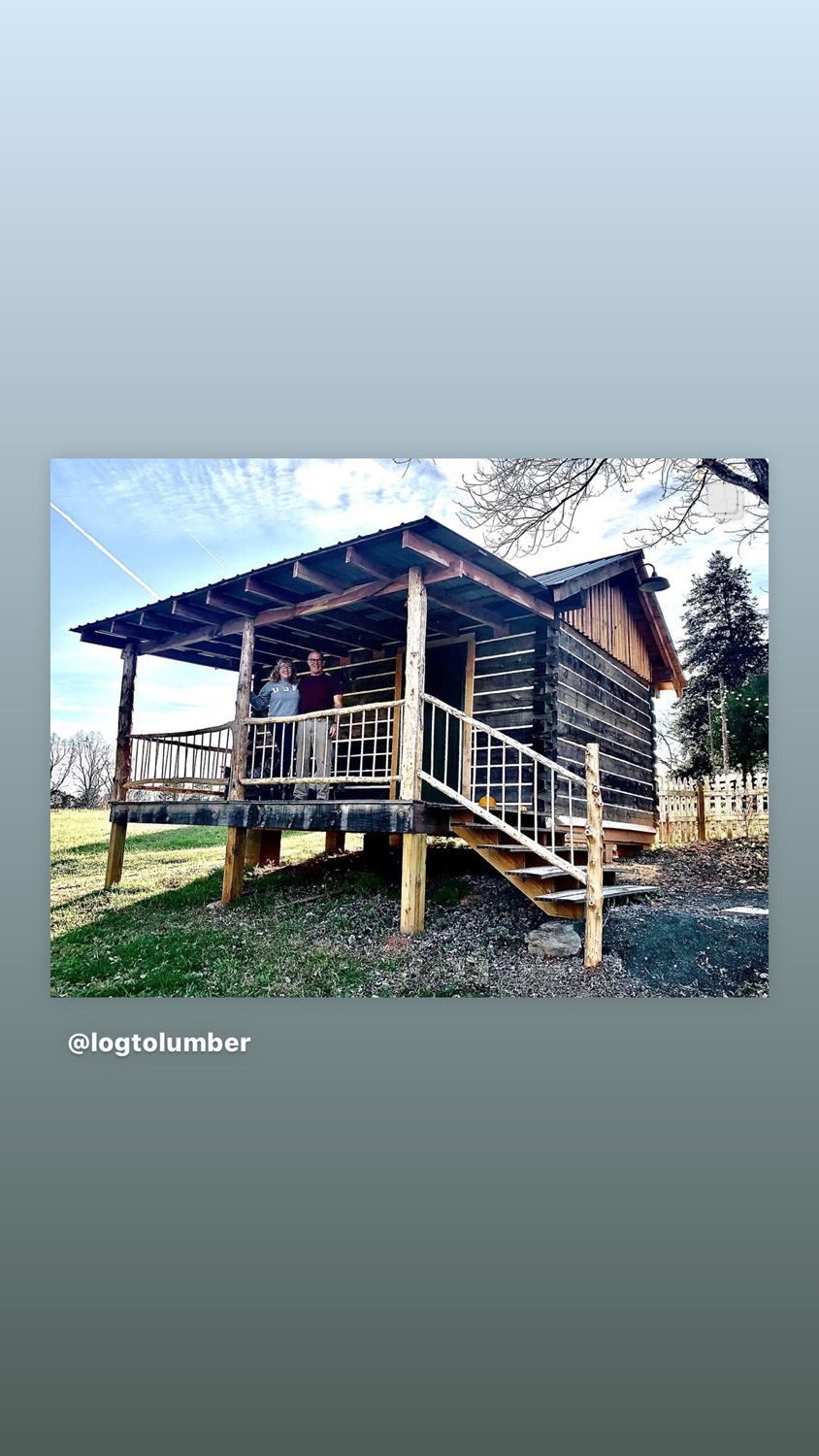 Rustic wooden cabin on stilts with a porch, set in a grassy rural area under a blue sky