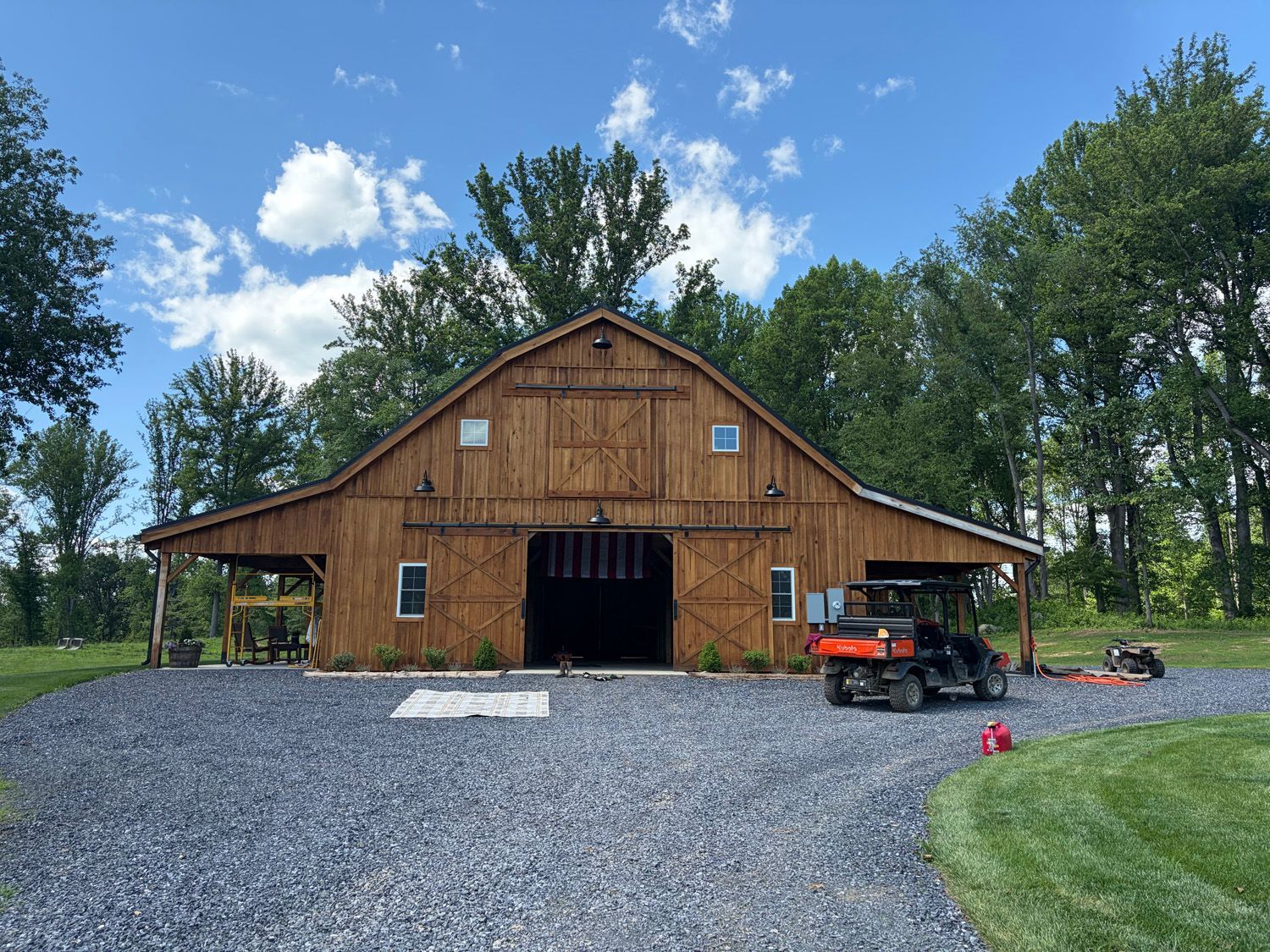 Brown barn with open doors on a gravel driveway, with an ATV parked beside it under a blue sky.