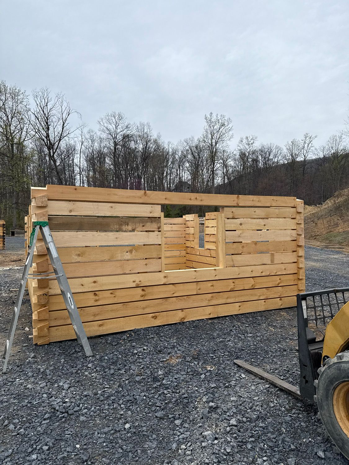 Wooden wall under construction on gravel, with a ladder and tractor tire visible outdoors.