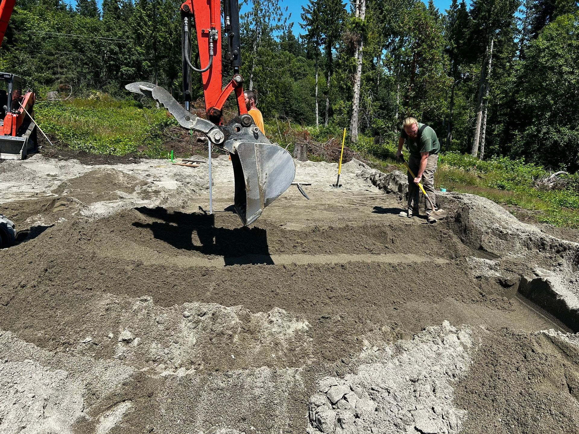 Construction site: excavator and worker leveling soil. Sunny day, forest background.