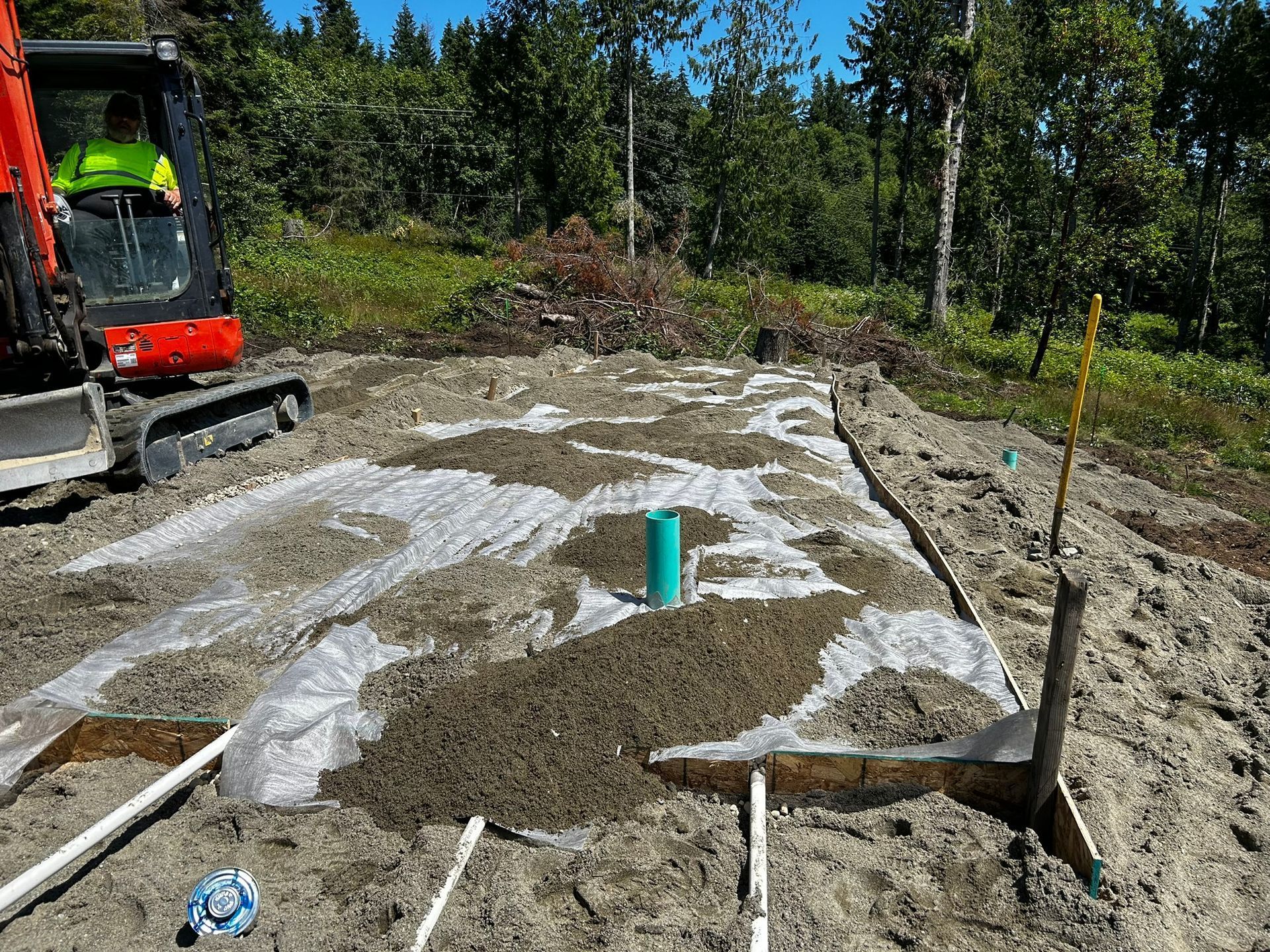 Construction site with mini excavator, plastic sheeting, sand, and PVC pipes.