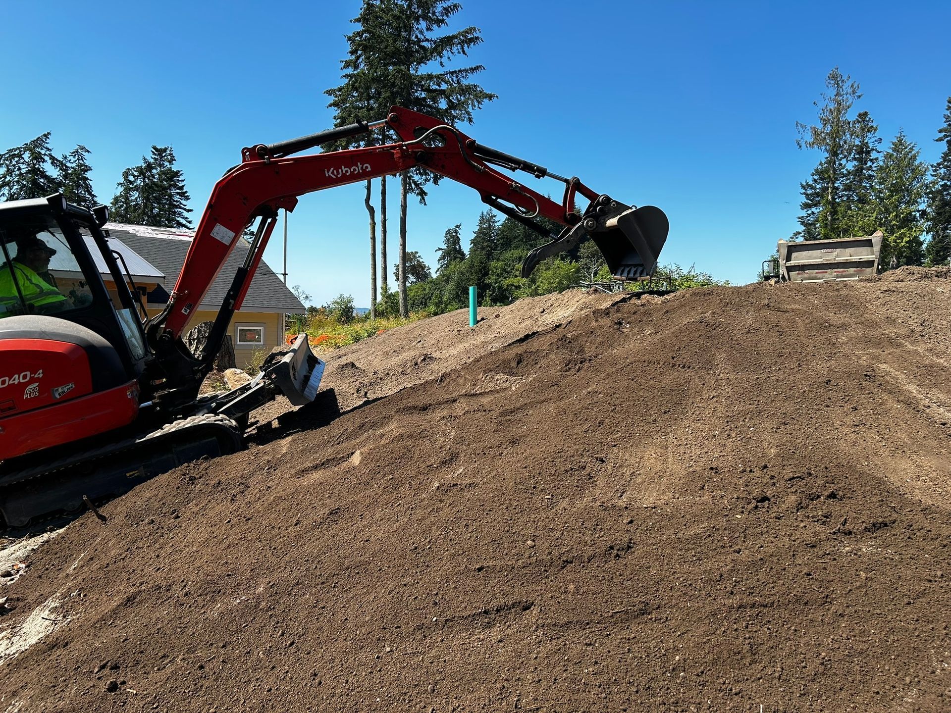 Red excavator on a brown hill, scooping soil. Bright sunny day with trees and a house in the background.