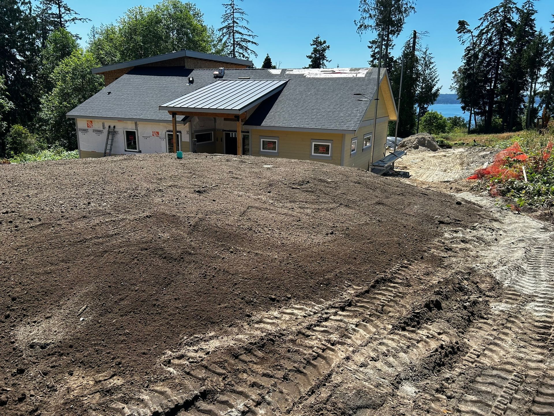 Newly built house with exposed exterior walls, sitting on a leveled dirt lot, next to water and trees.