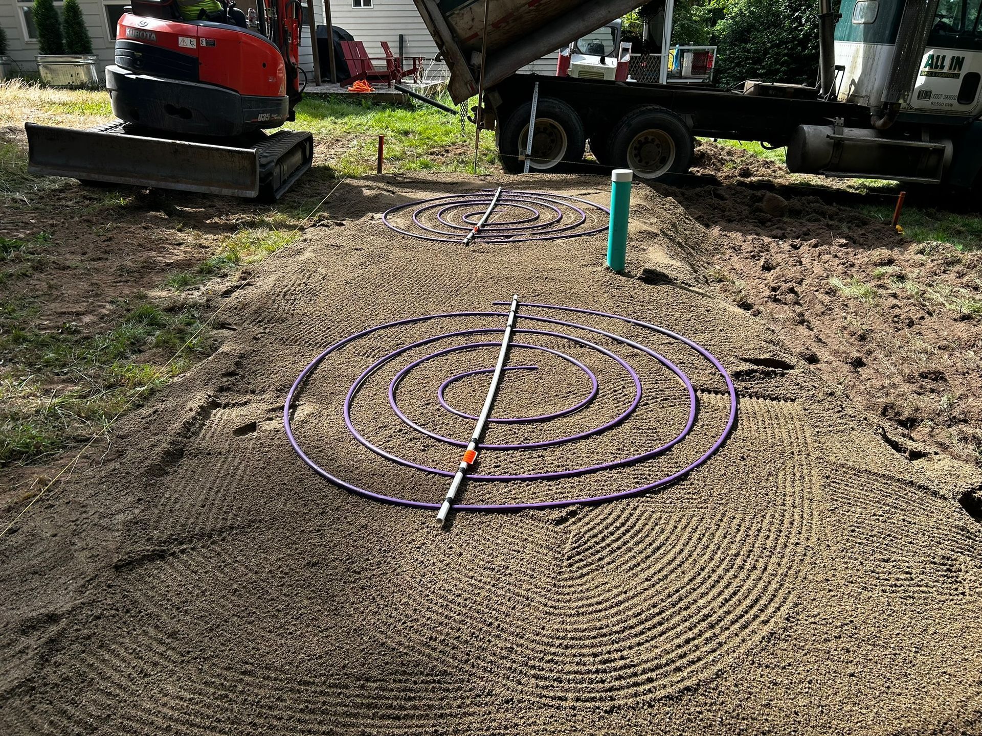 Construction site with purple pipes arranged in concentric circles on a sandy surface. An excavator and truck are visible.