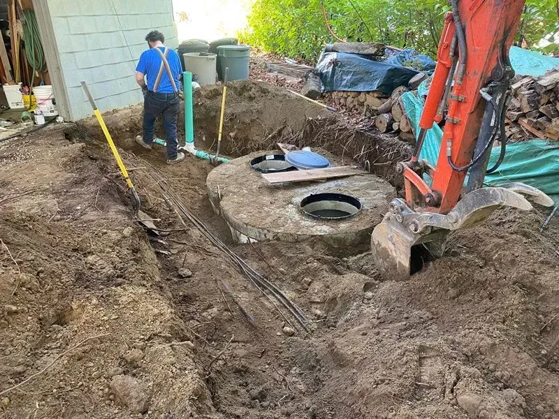 Excavation site with a septic tank; worker in blue suspenders near piping, excavator at right.