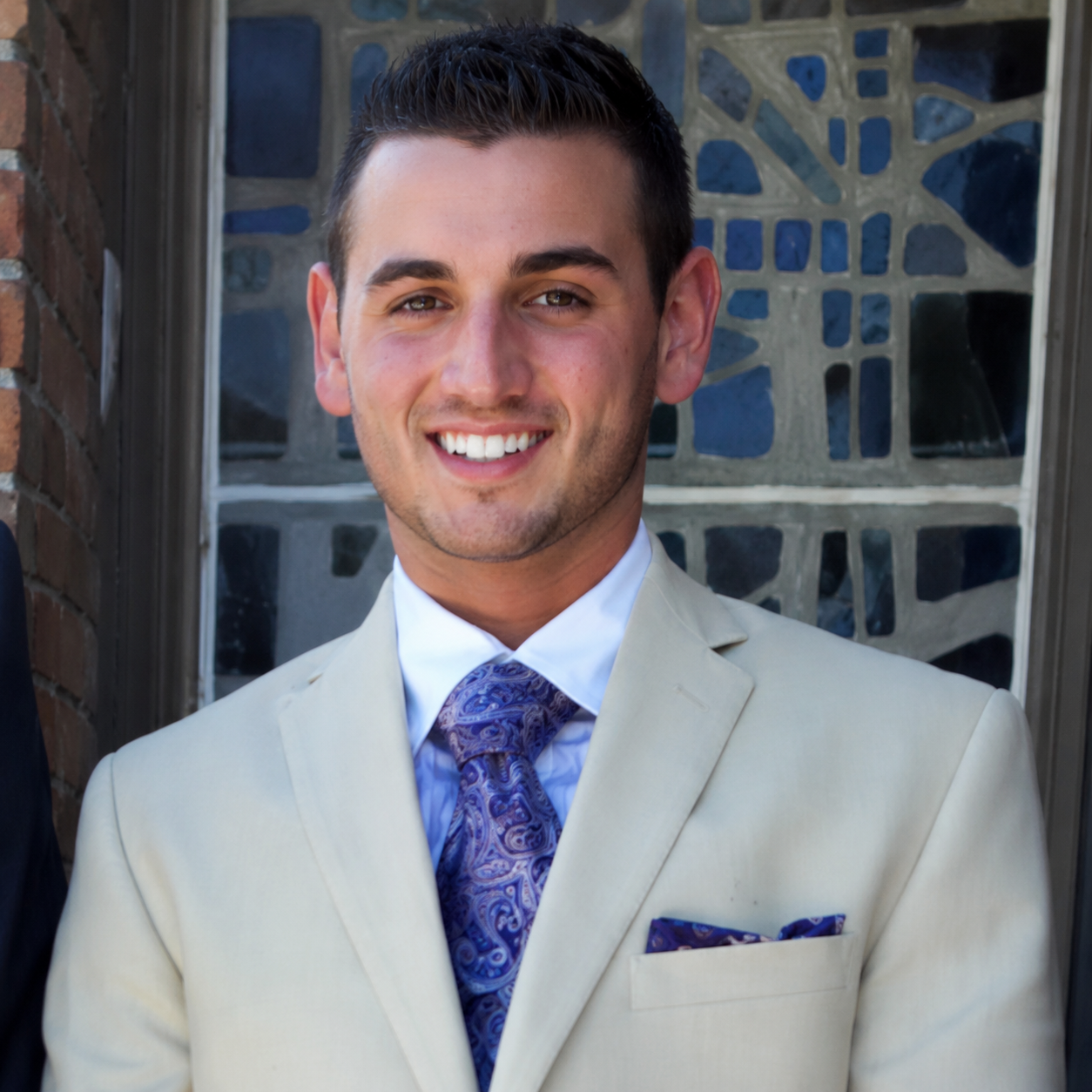Smiling man in a light suit with a blue tie and pocket square, standing before a stone wall.