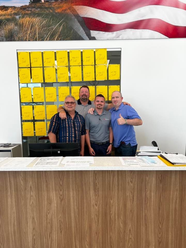 Four people posing behind a front desk in an office, with yellow notes on the wall and an American flag above.