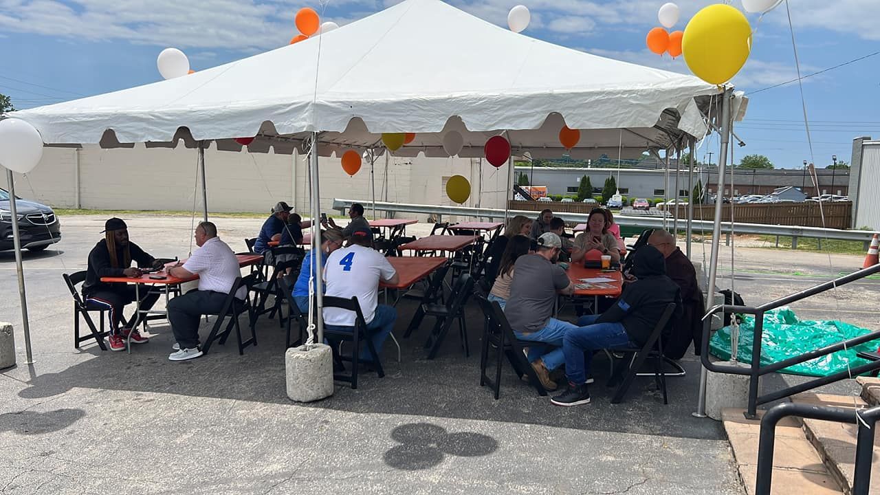 People seated under a white canopy at an outdoor gathering with balloons and tables on a sunny day