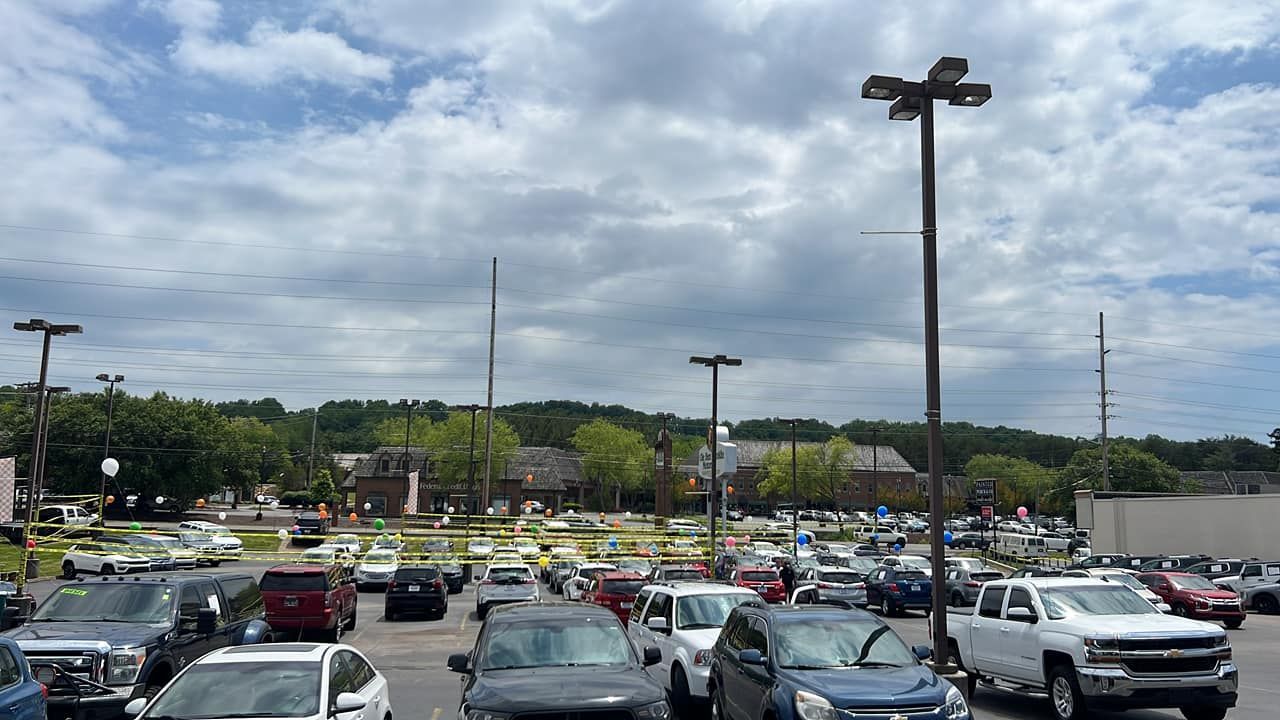 Parking lot filled with cars under cloudy sky, with trees and tall light poles in the background