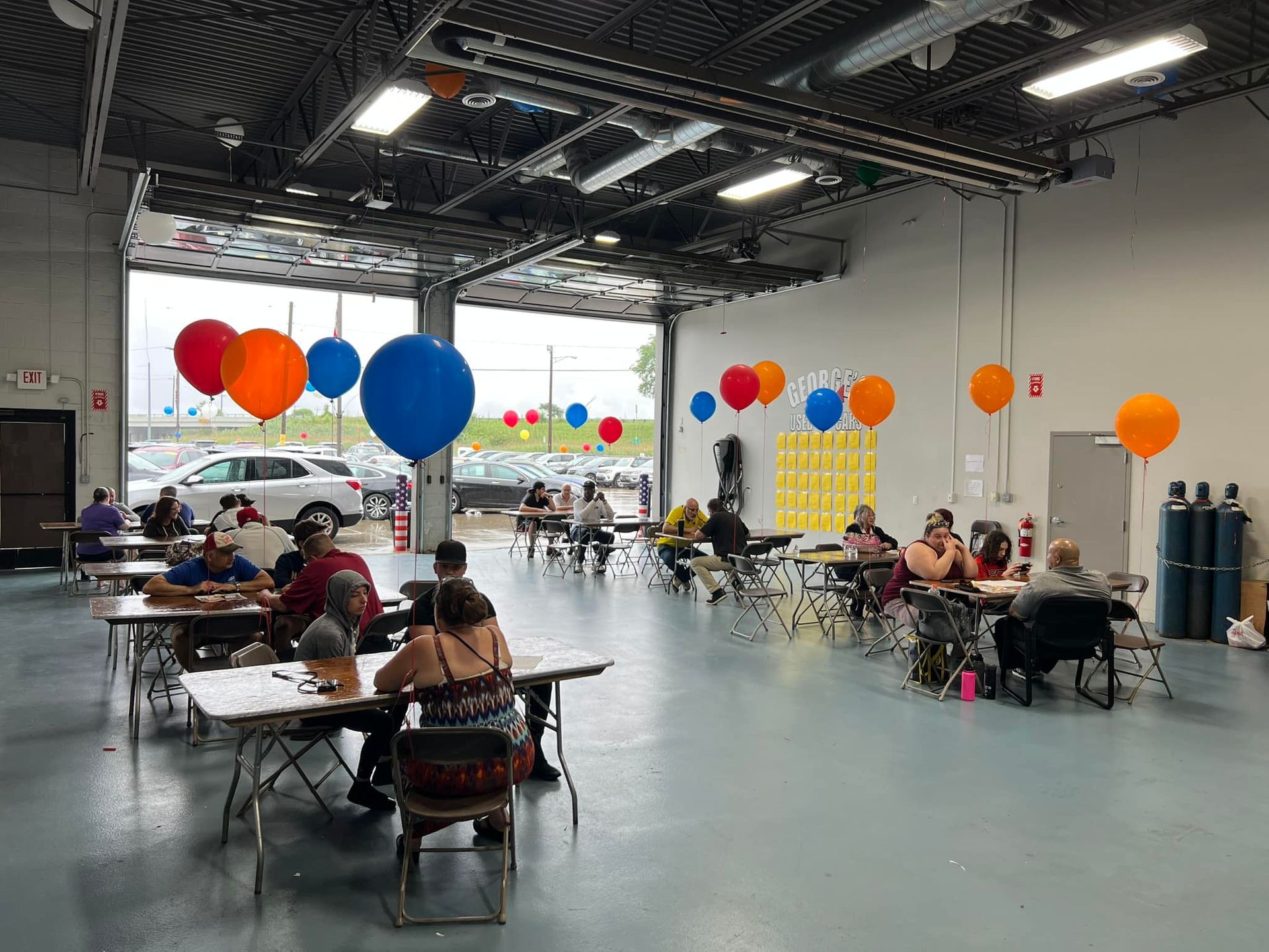 People seated at tables in a large decorated hall with colorful balloons and bright overhead lights