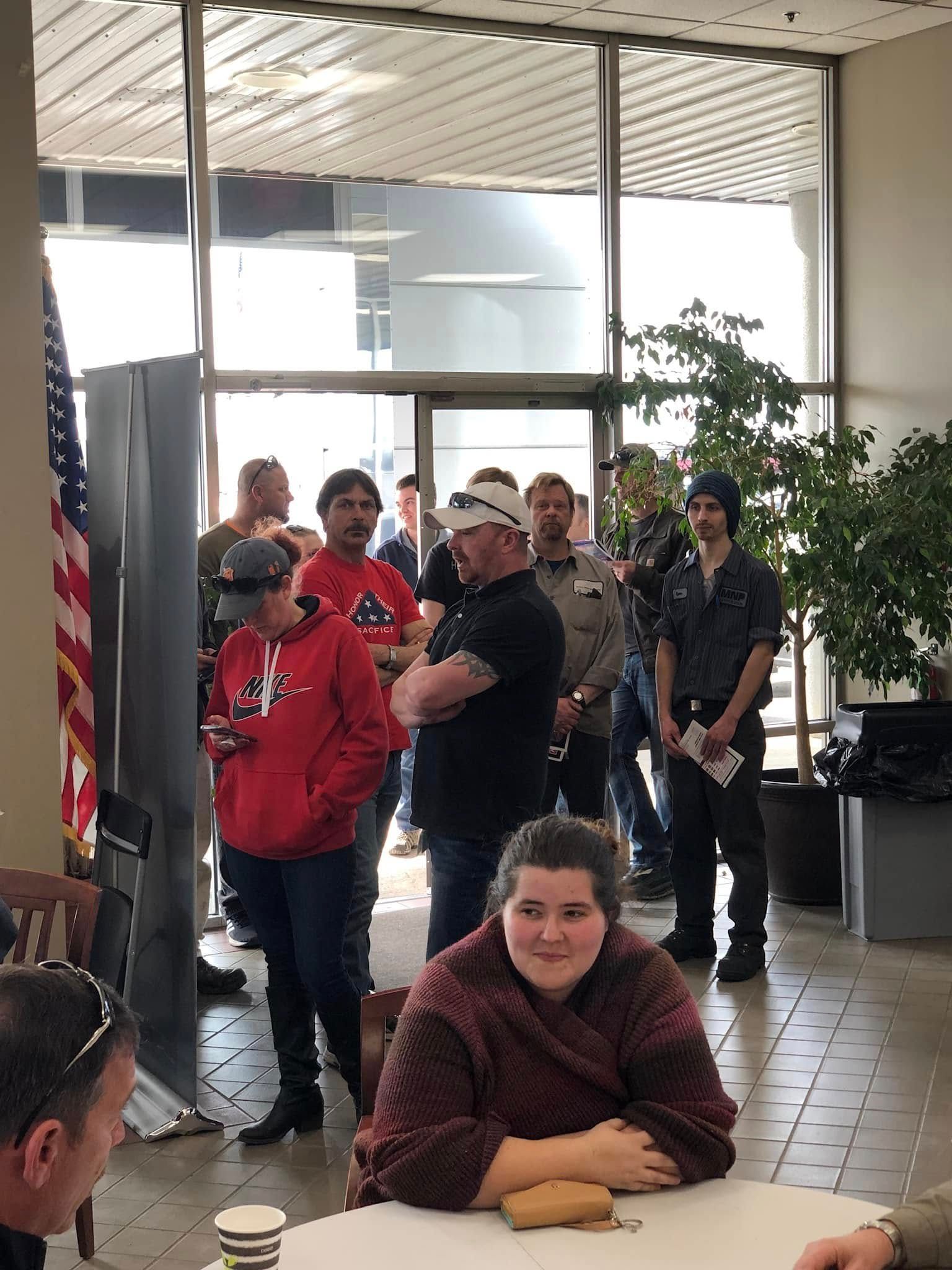 People waiting in a lobby, with a seated woman in the foreground and others standing by the glass doors.