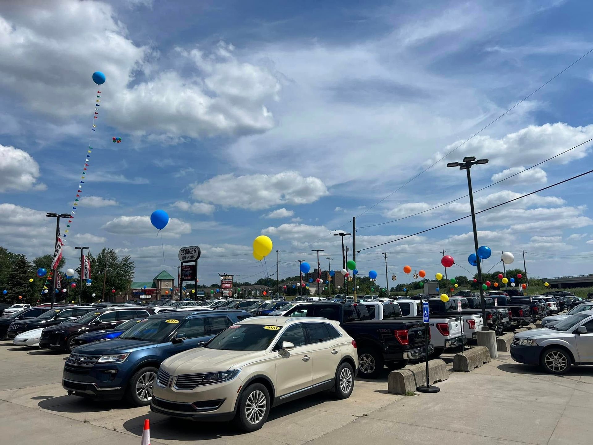 Outdoor car lot with rows of parked vehicles and colorful balloons under a cloudy sky