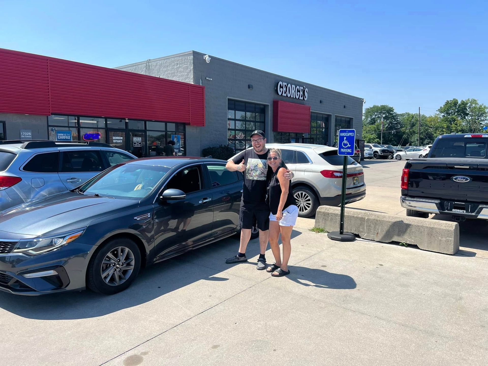 Two people standing beside a black car in a sunny parking lot outside a store