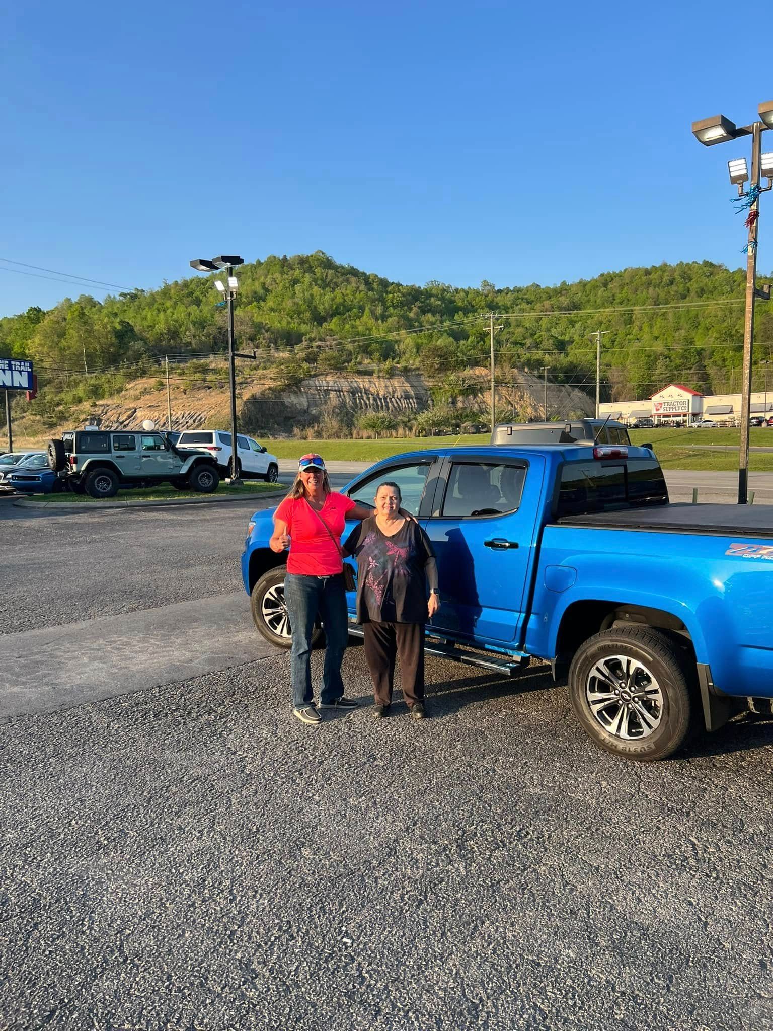 Two people standing beside a blue pickup truck on a gravel lot with hills and clear sky behind them