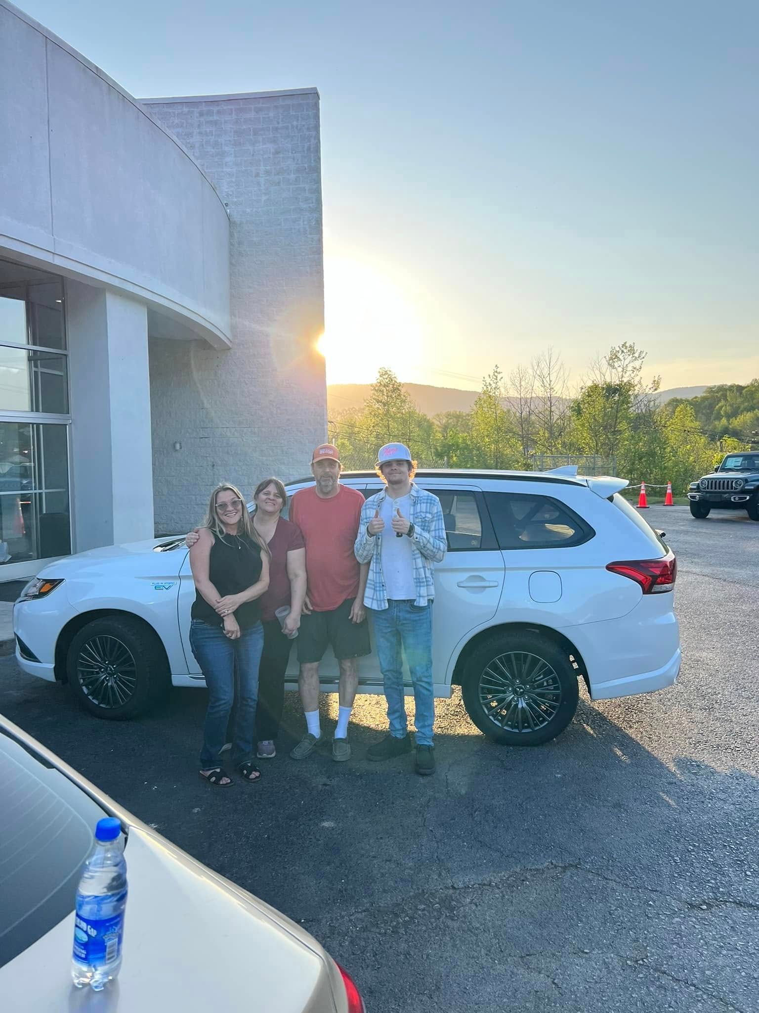 Three people standing beside a white SUV in a parking lot at sunset