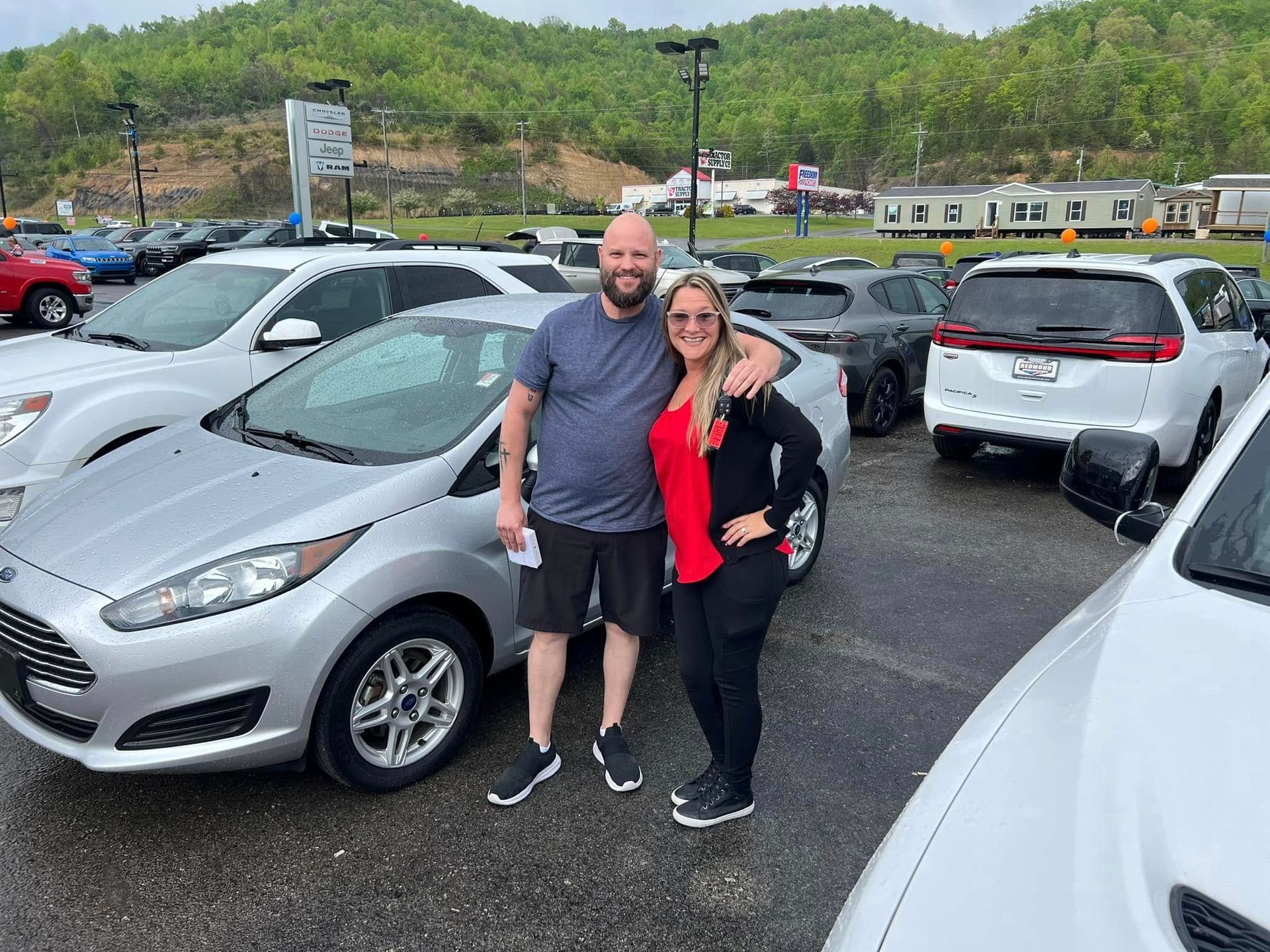 Two people posing in a parking lot beside a silver car, with green hills in the background.