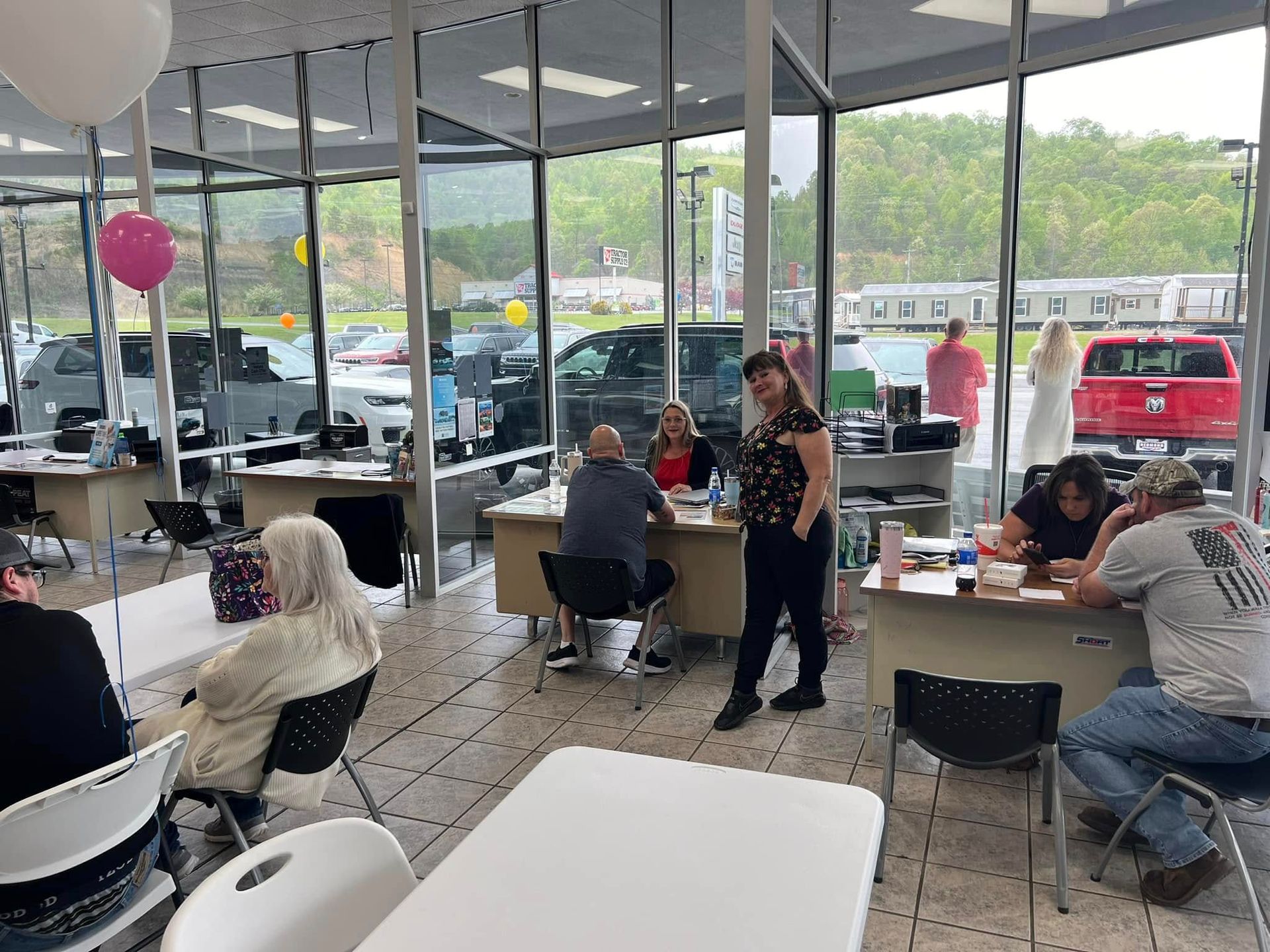 People seated at tables in a bright glass-walled café, with balloons and parked cars visible outside.