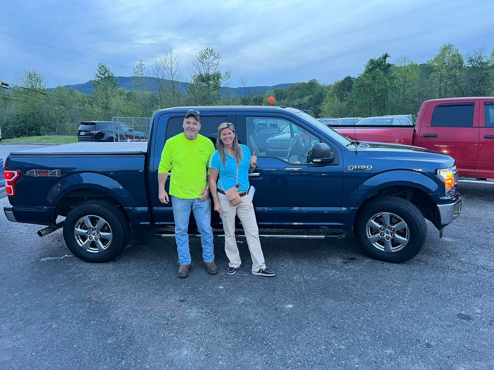 Two people standing beside a dark blue pickup truck in a gravel lot, with trees and other vehicles in the background