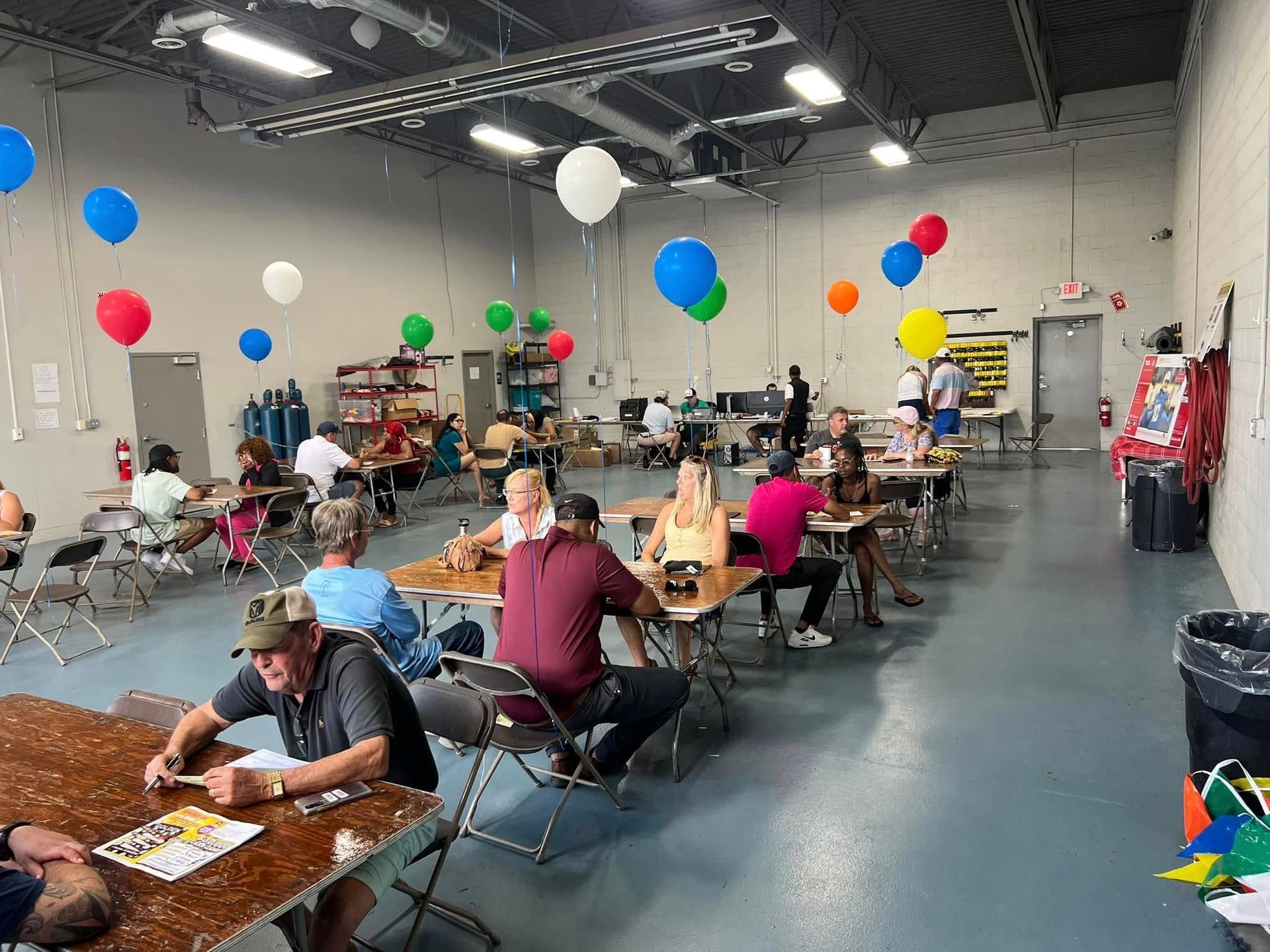People seated at tables in a decorated indoor hall with colorful balloons and overhead lights