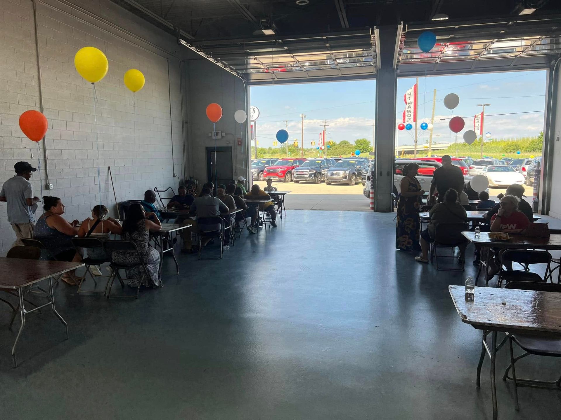 Indoor dining hall with people seated at tables, colorful balloons on the walls, and an open view to the outside.