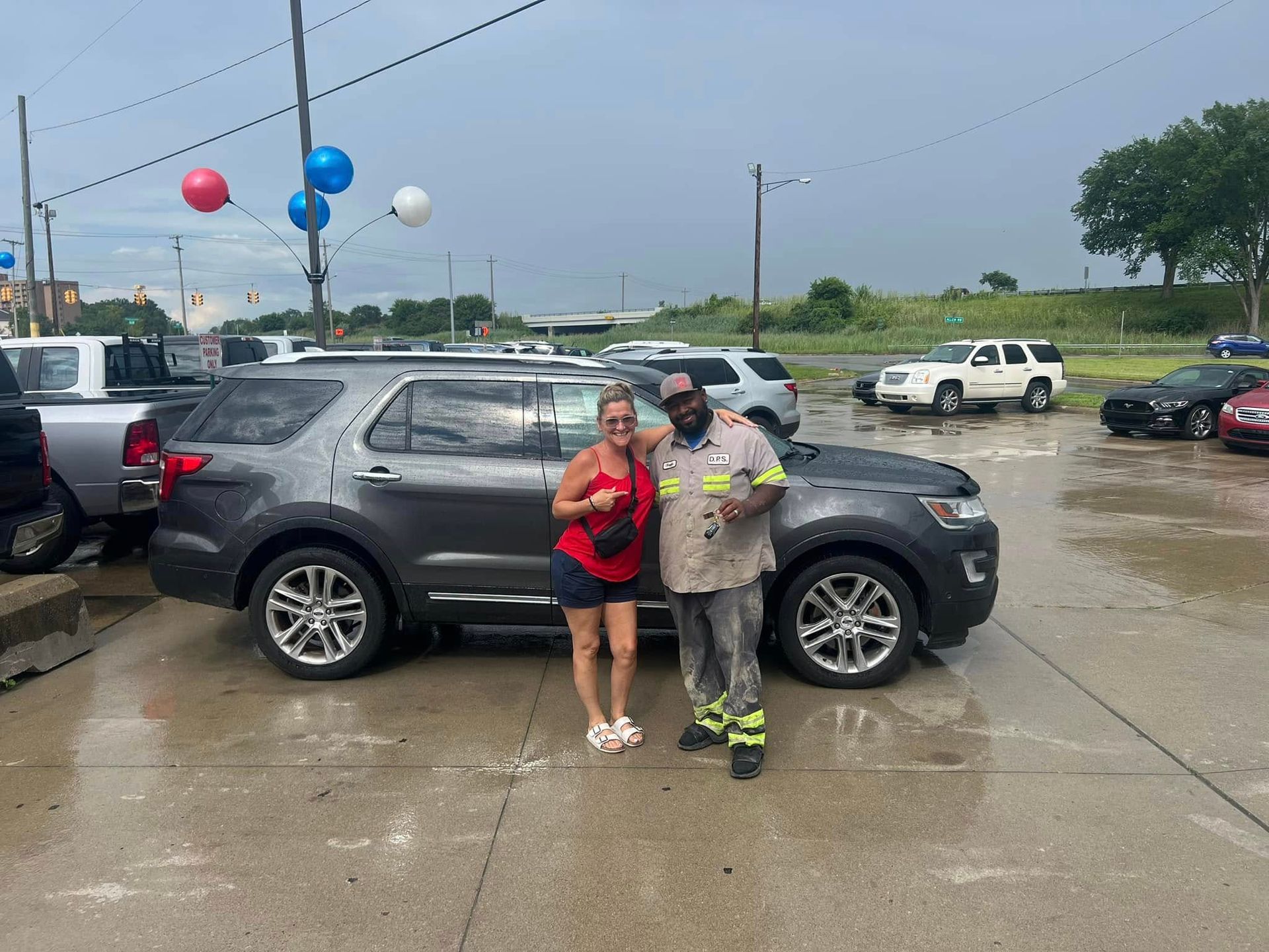 Two people pose beside a black SUV in a wet parking lot with balloons overhead.