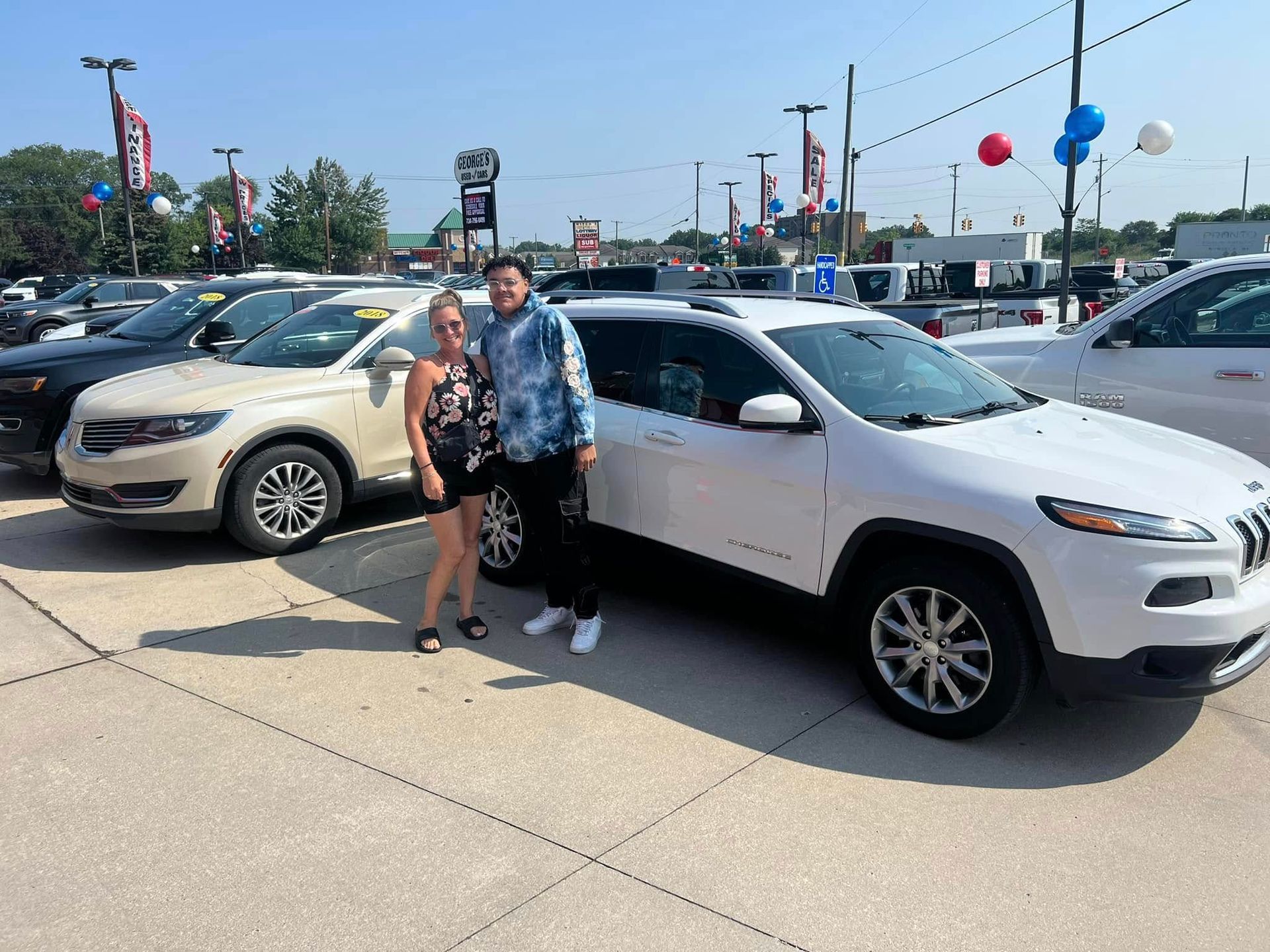 Two people standing beside a white SUV in a car dealership lot on a sunny day